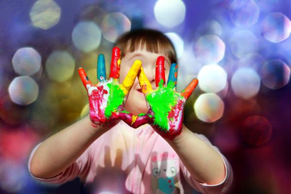 smiling young child with colourfully painted hands in front of her face