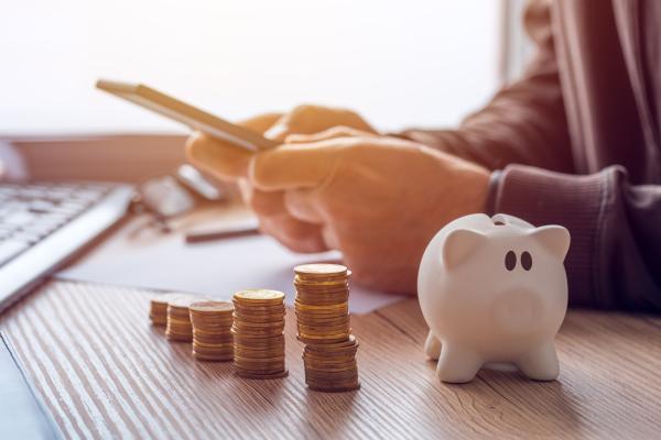 A person's hands holding a phone, next to piles of coins and a piggy bank