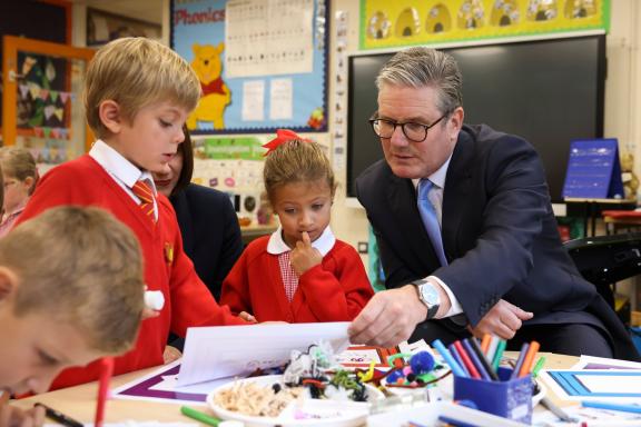 Man in a suit (Prime Minister) speaks with two young pupils at a table in a classroom